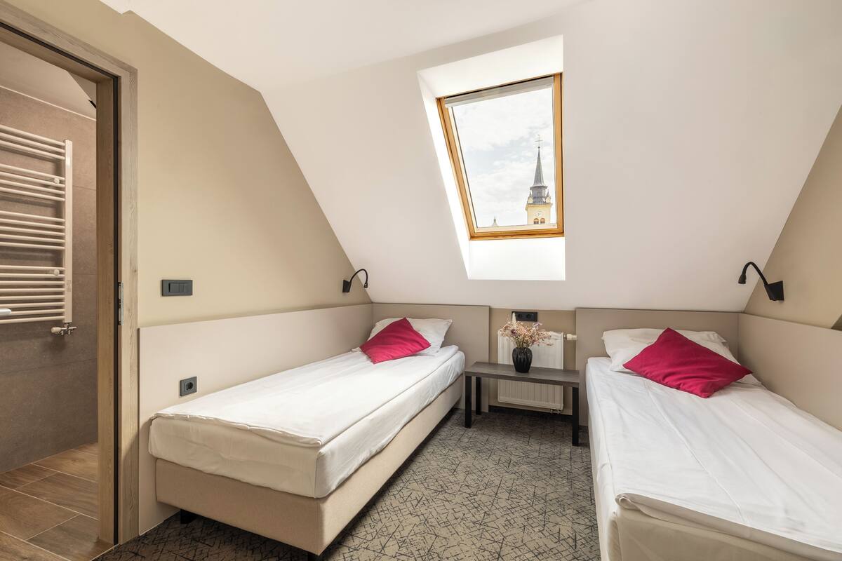 Una habitación pequeña y moderna con dos camas individuales, una mesita y una ventana con vistas a la aguja de una iglesia.