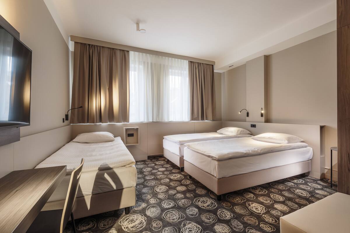 Habitación de hotel con dos camas individuales, un escritorio, una silla, un televisor y un ventanal grande con cortinas.