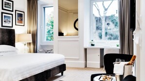 A hotel room with a large bed, a small table with a tray of desserts, a chair, and a view of greenery through the window. - The Shire Hotel (Rome)