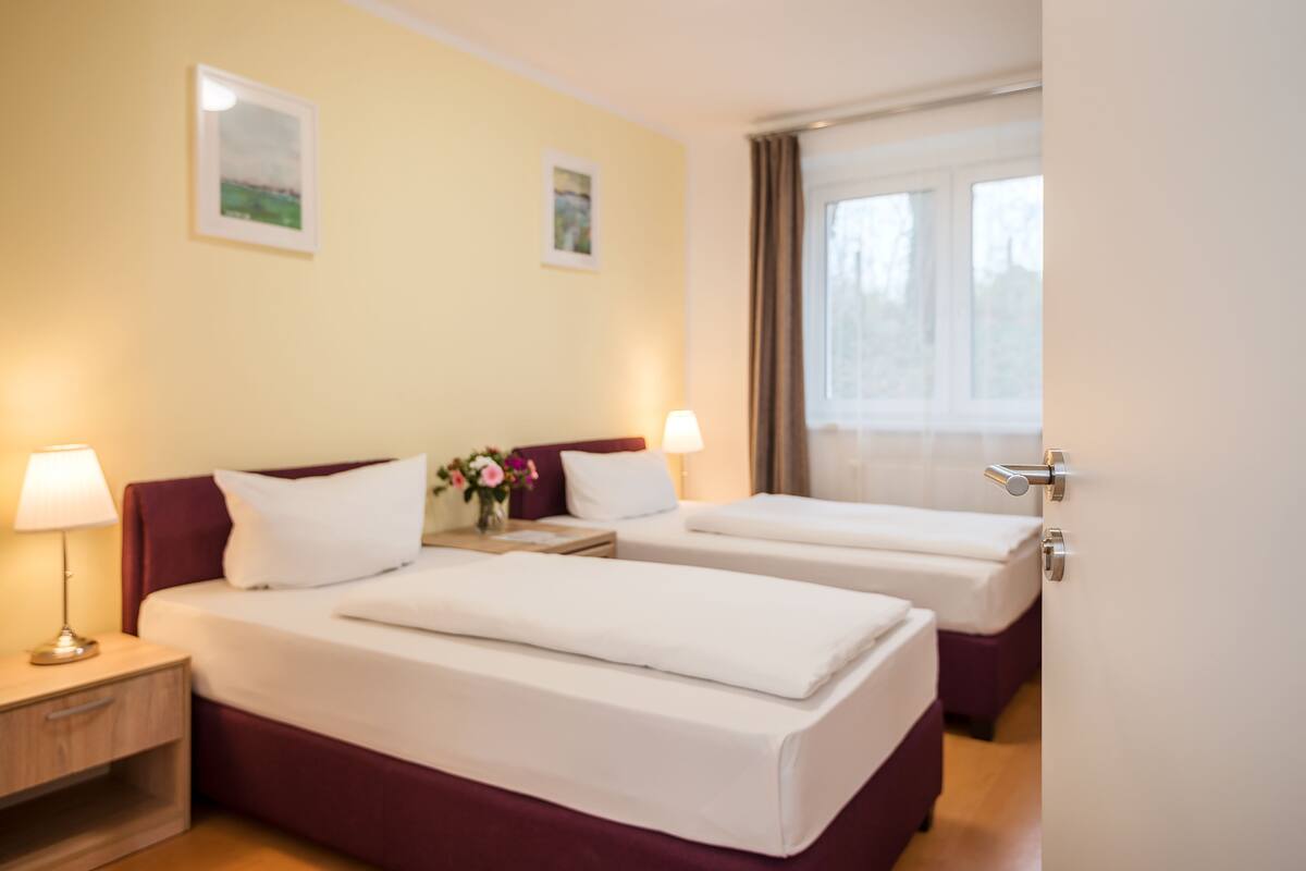 Habitación de hotel con dos camas, una mesita de noche con lámpara y una ventana con cortinas.