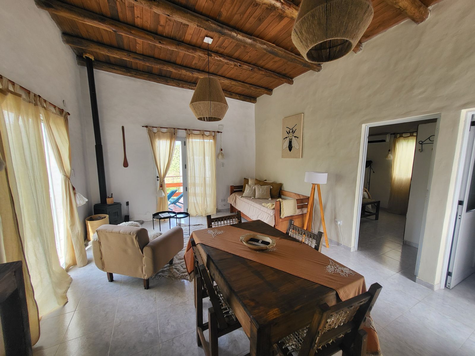 A cozy room with a wooden dining table, a sofa, and a bed. There's a window with curtains, a lamp, and a wall-mounted insect art piece.