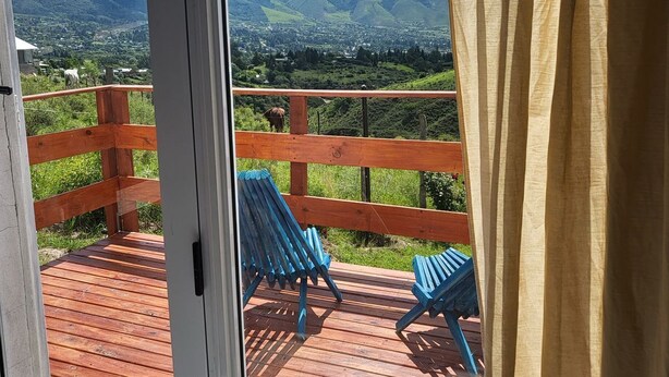 A wooden deck with blue chairs, a wooden railing, and a view of green hills and mountains.
