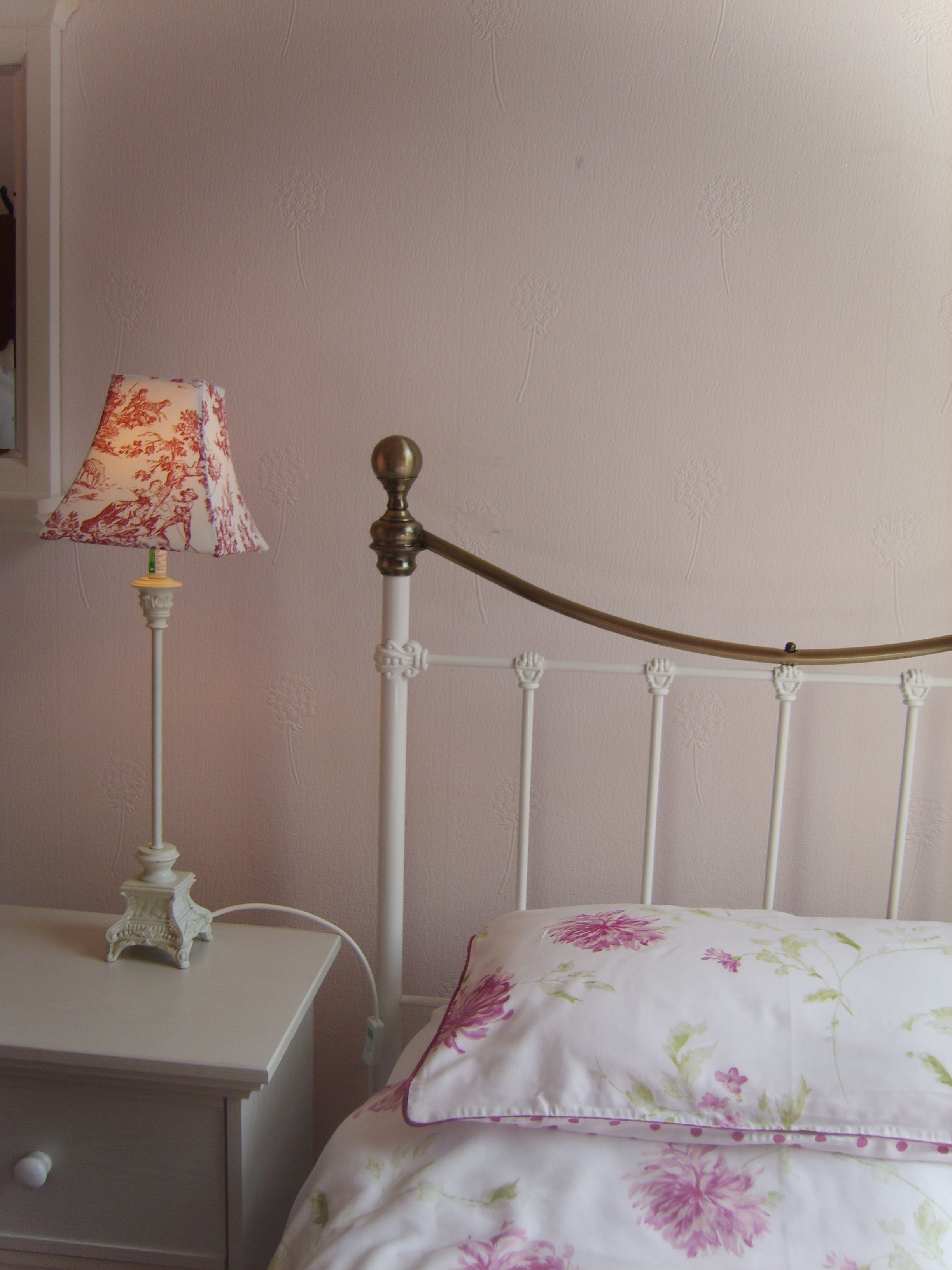 A bedroom with a white bed, floral bedding, a nightstand with a lamp, and a white wall.