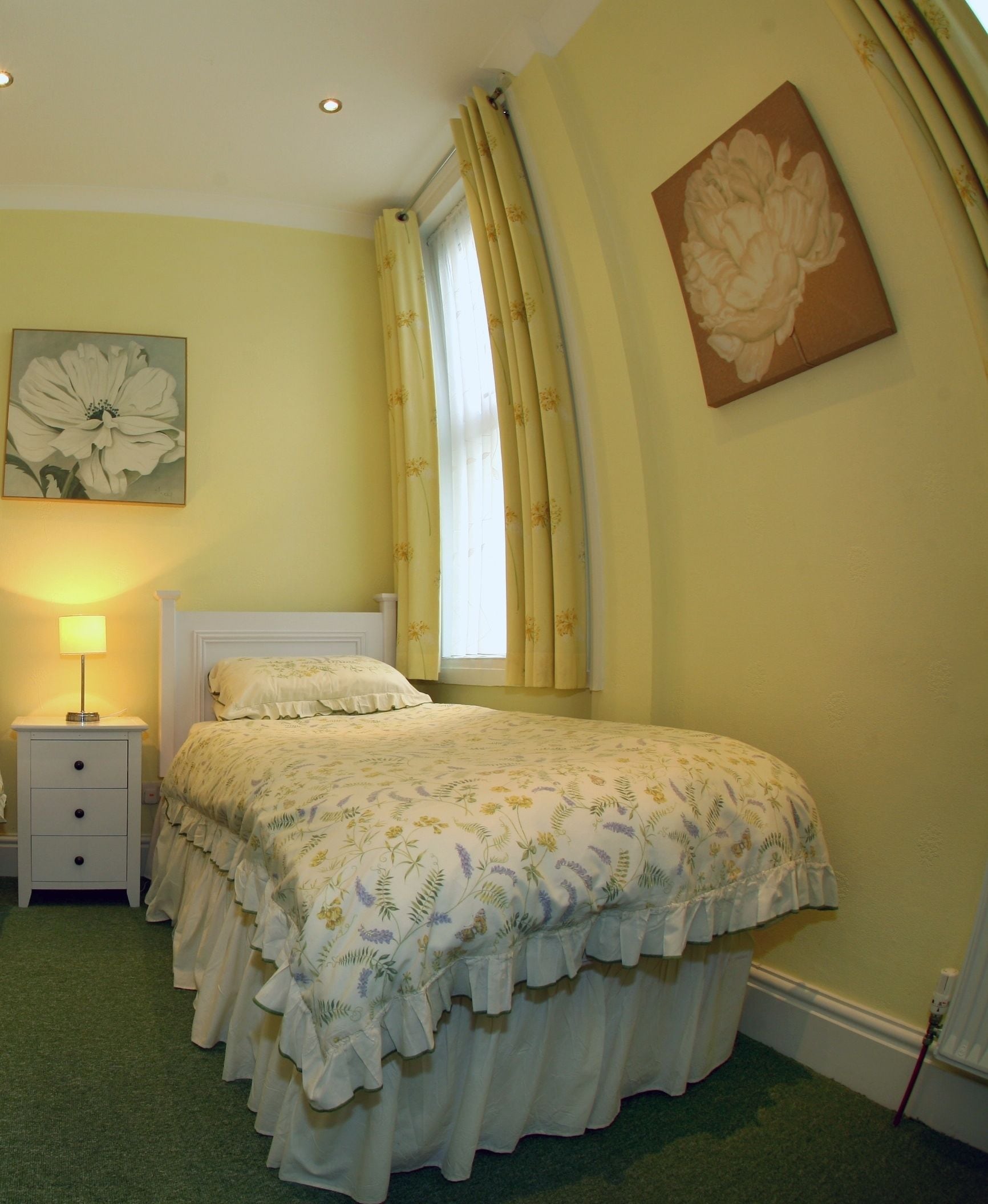 Un dormitorio con cama, mesita de noche, lámpara, ventana con cortinas y un cuadro con motivos florales en la pared.