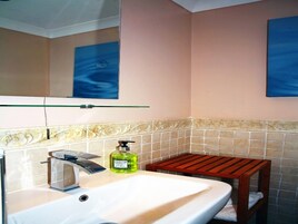 Un baño con un lavabo blanco, un espejo, una botella verde de jabón de manos y un taburete de madera.