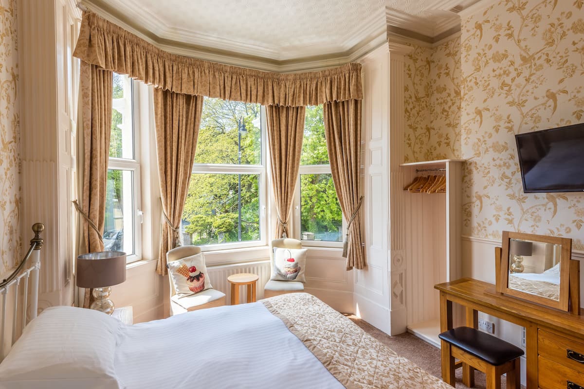 Une chambre d’hôtel avec un grand lit, une commode en bois, un miroir, une chaise et une fenêtre avec des rideaux.