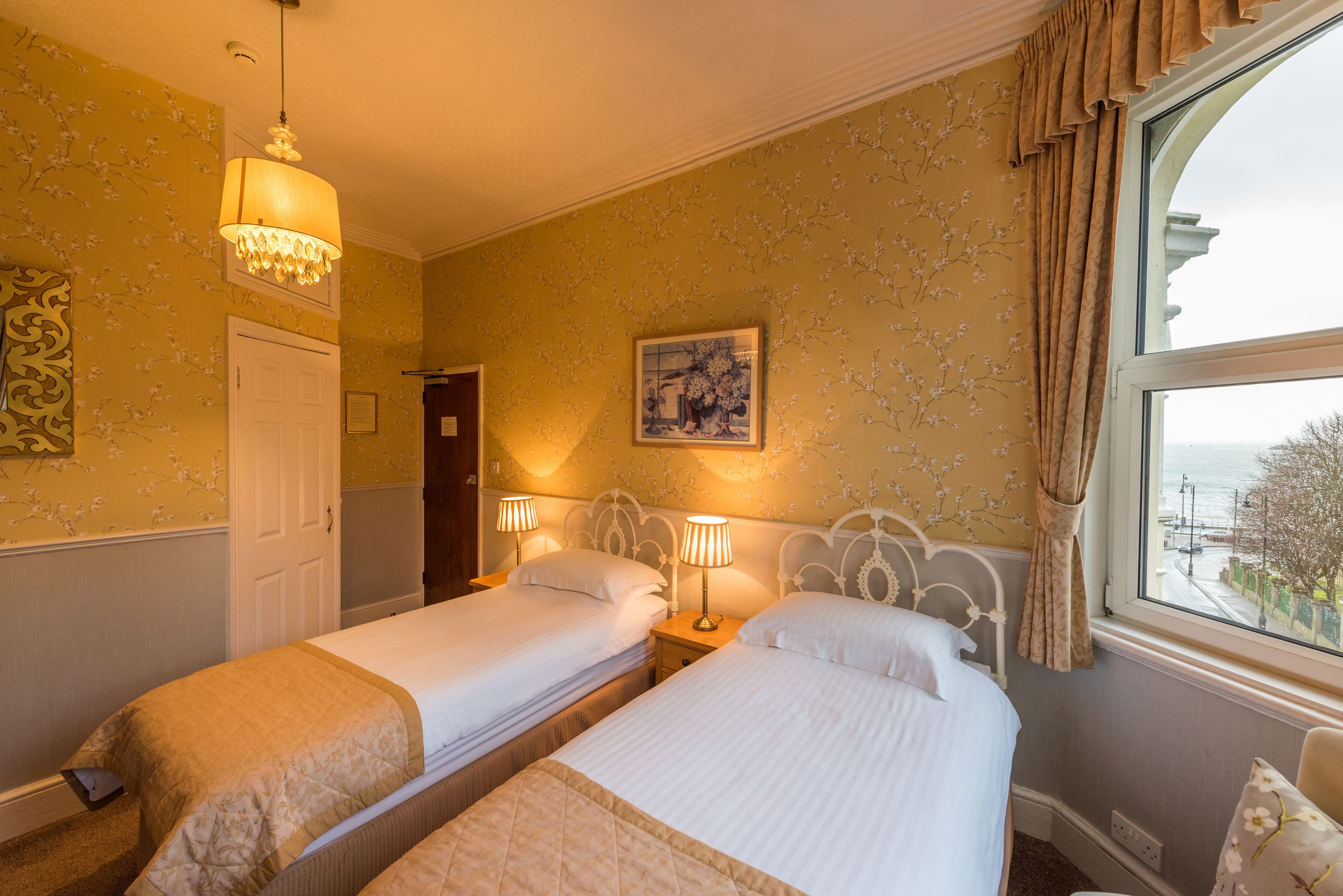 A hotel room with two beds, a chandelier, a painting, and a window with a view.