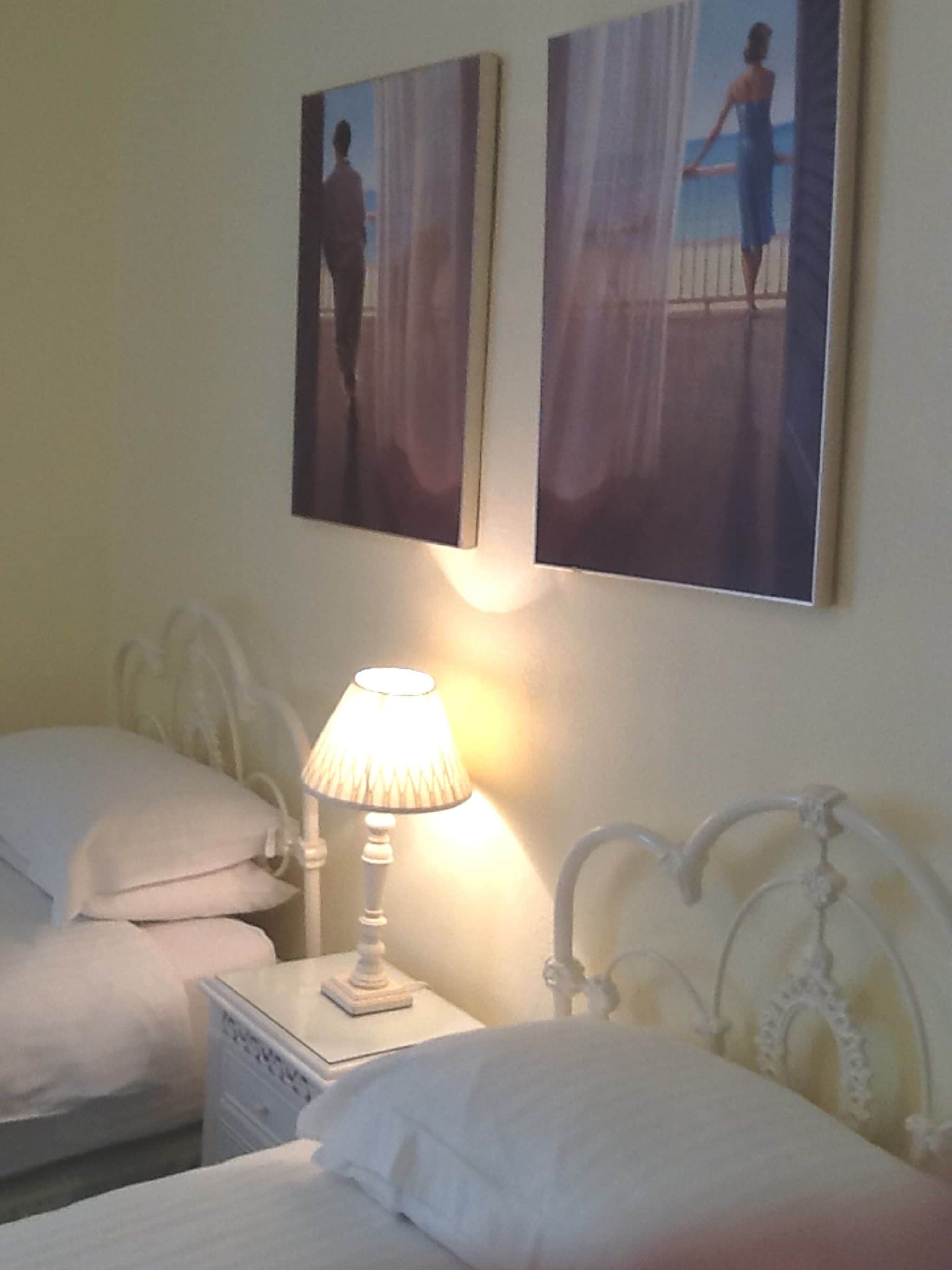 A hotel room with two beds, a nightstand with a lamp, and two paintings on the wall.