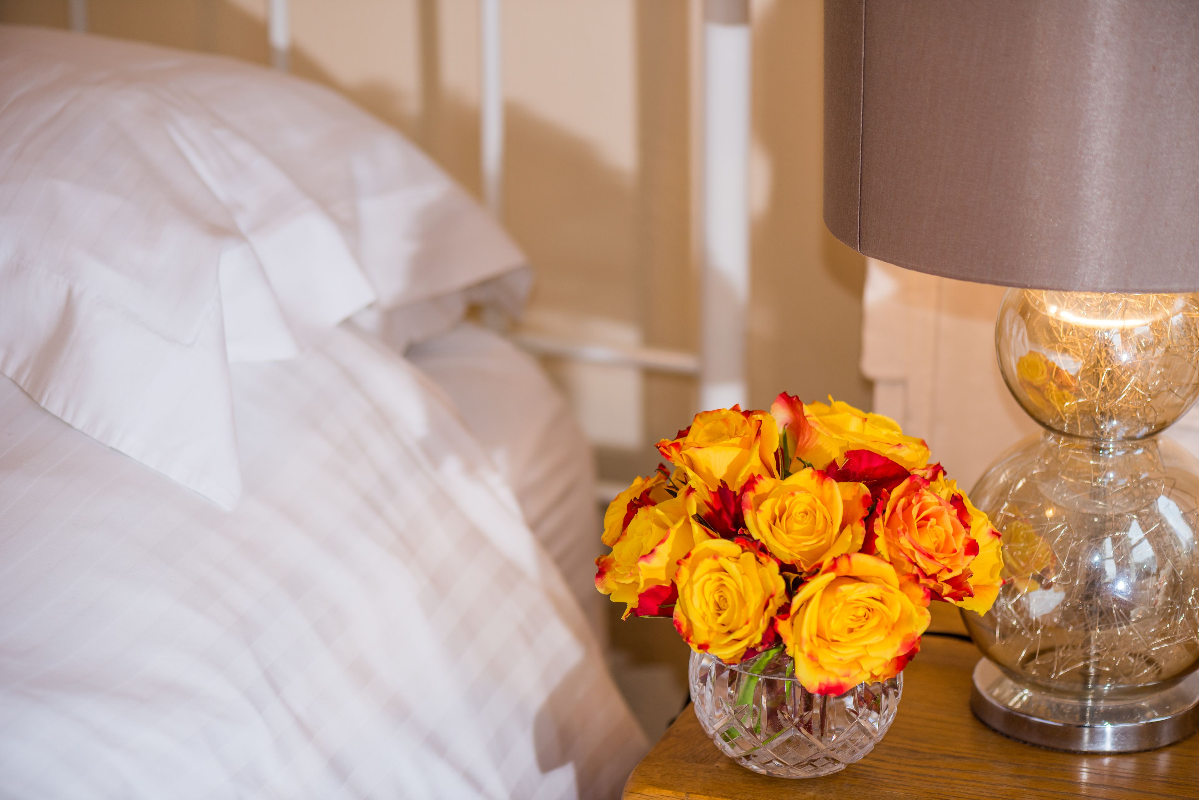 Un lit avec une literie blanche, un vase en verre transparent contenant des roses jaunes et une lampe posée sur une table de chevet en bois.