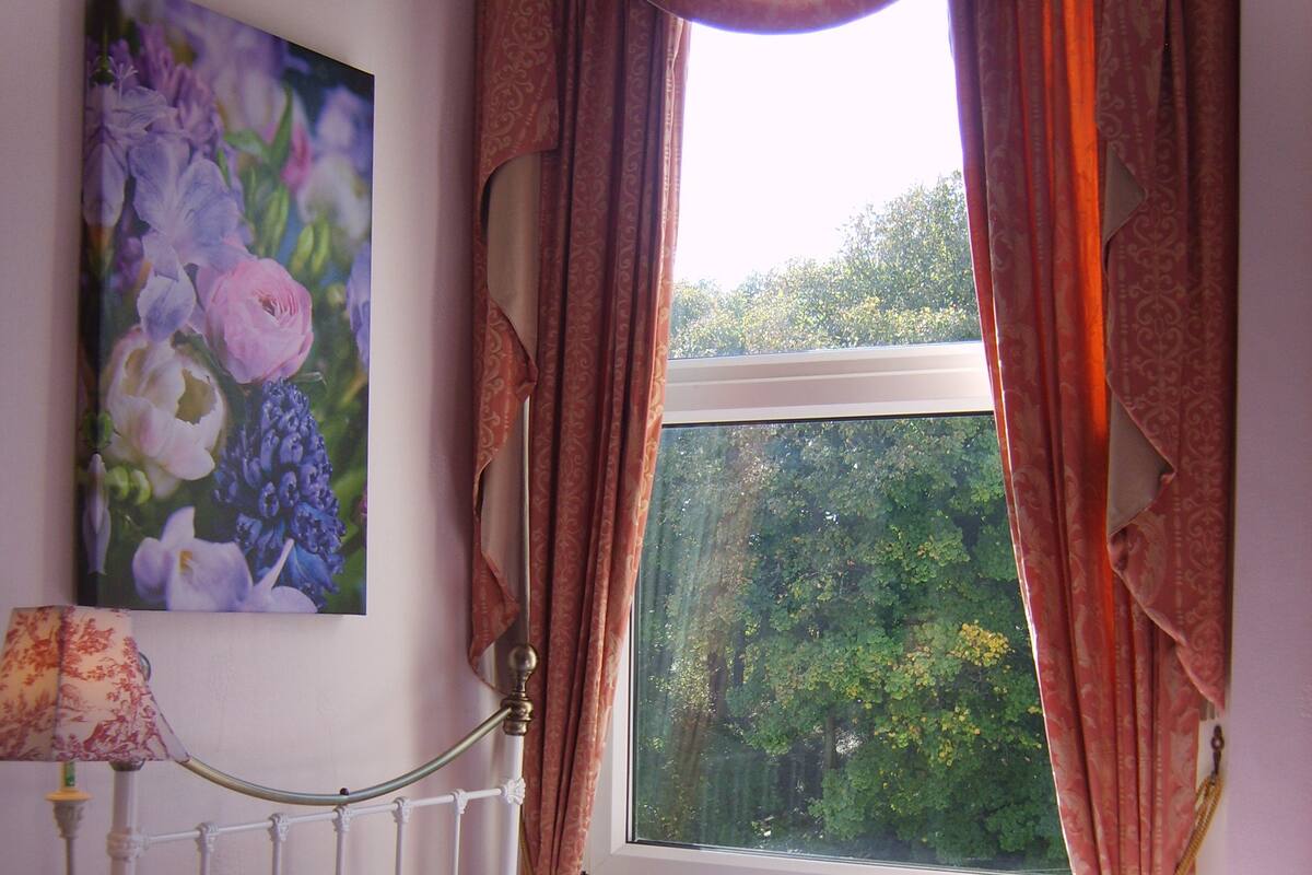 Une chambre à coucher avec un lit, une table de chevet, une lampe, une fenêtre avec des rideaux et un tableau au mur.