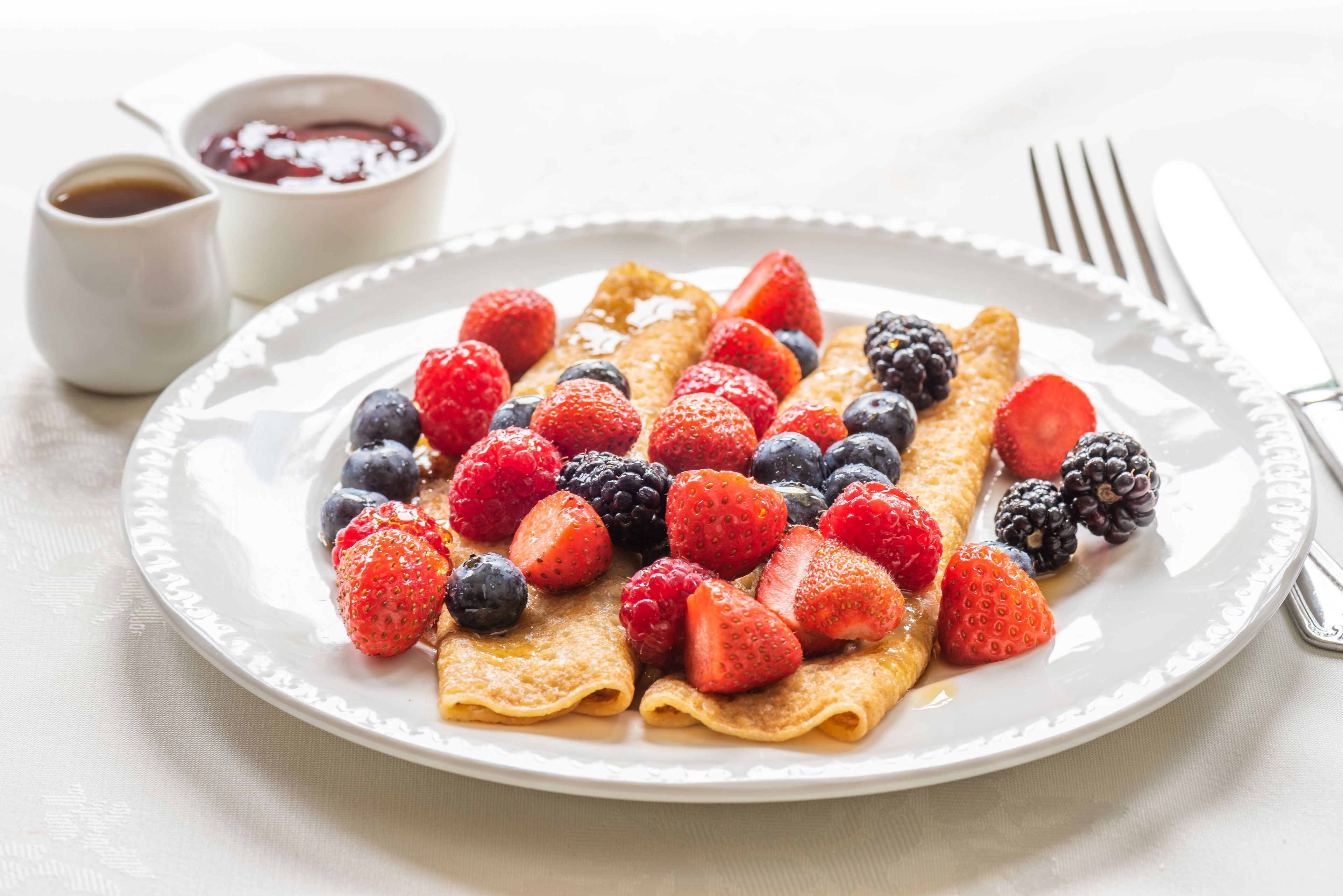 A plate of crepes topped with strawberries, blueberries, and blackberries, accompanied by a small bowl of syrup.