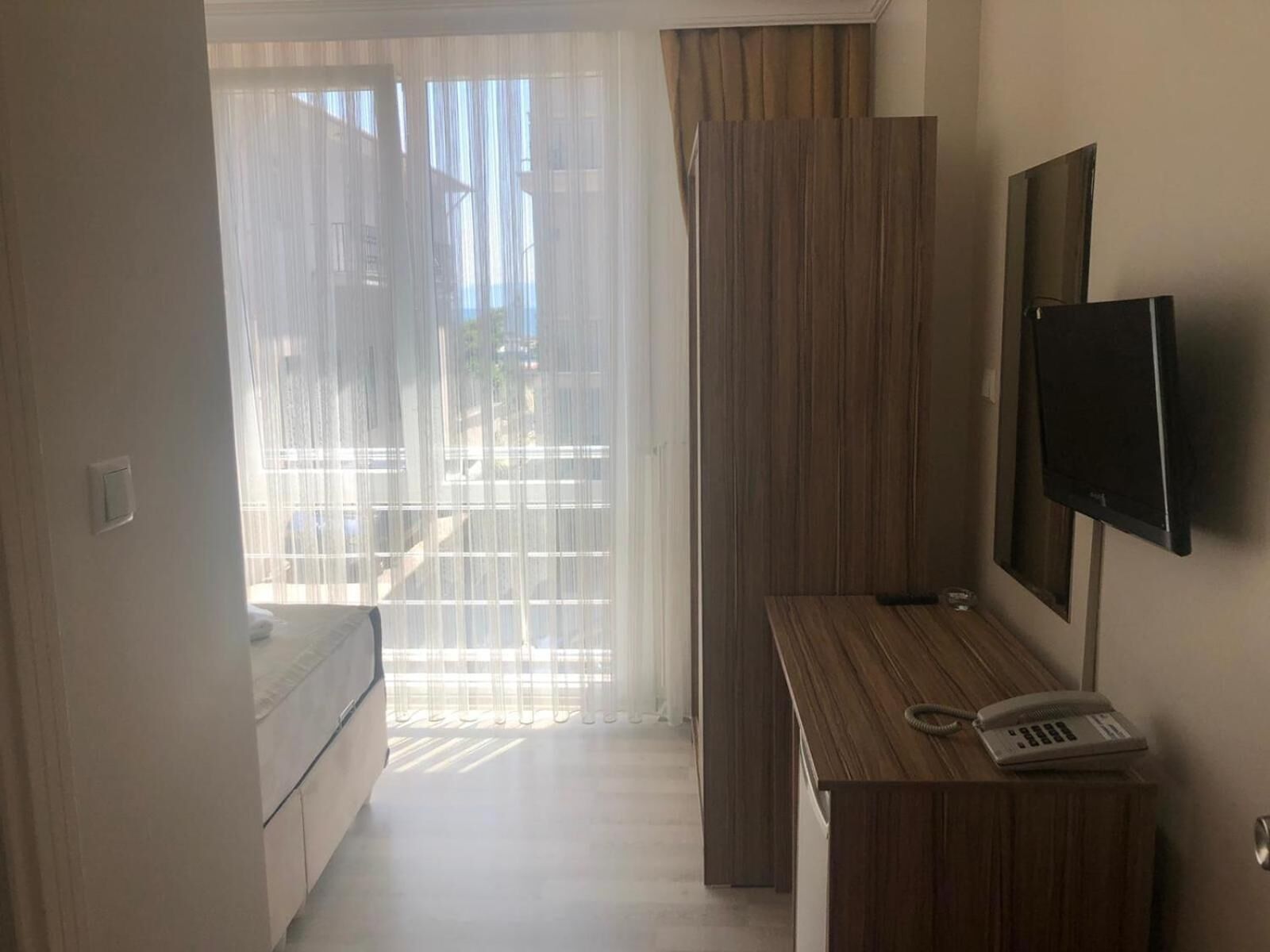 Een hotelkamer met een bed, een bureau met telefoon, een aan de muur bevestigde televisie en een raam met dunne gordijnen.