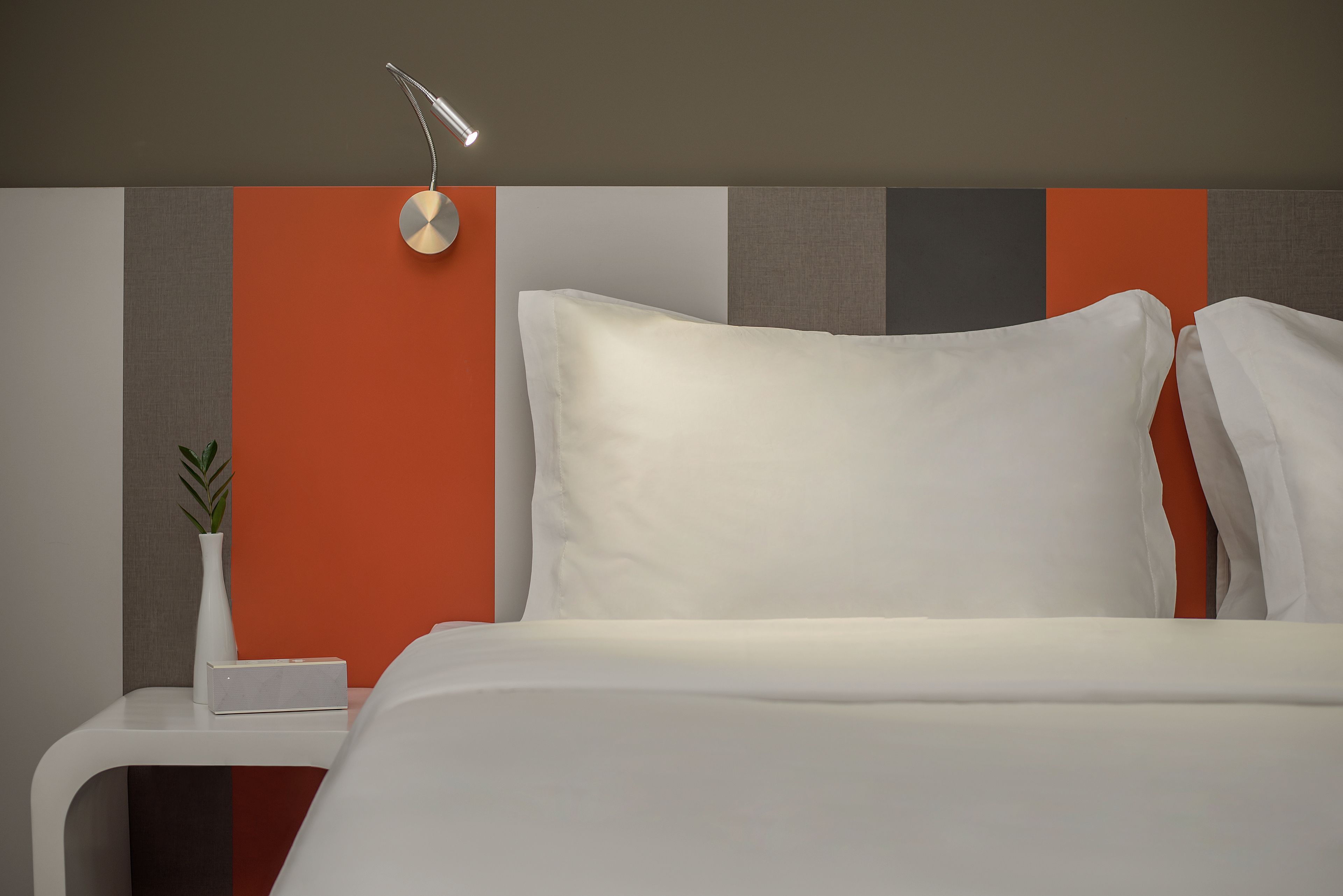 A neatly made bed with white bedding, a bedside table with a white vase and a small plant, and a modern wall with a mix of grey and orange panels.