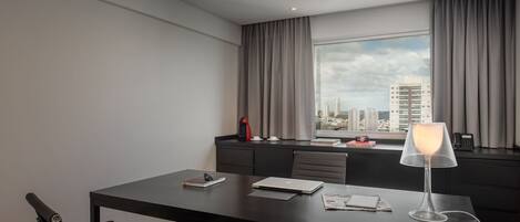 A modern hotel room with a large window, a desk with a lamp, and a view of the city skyline.