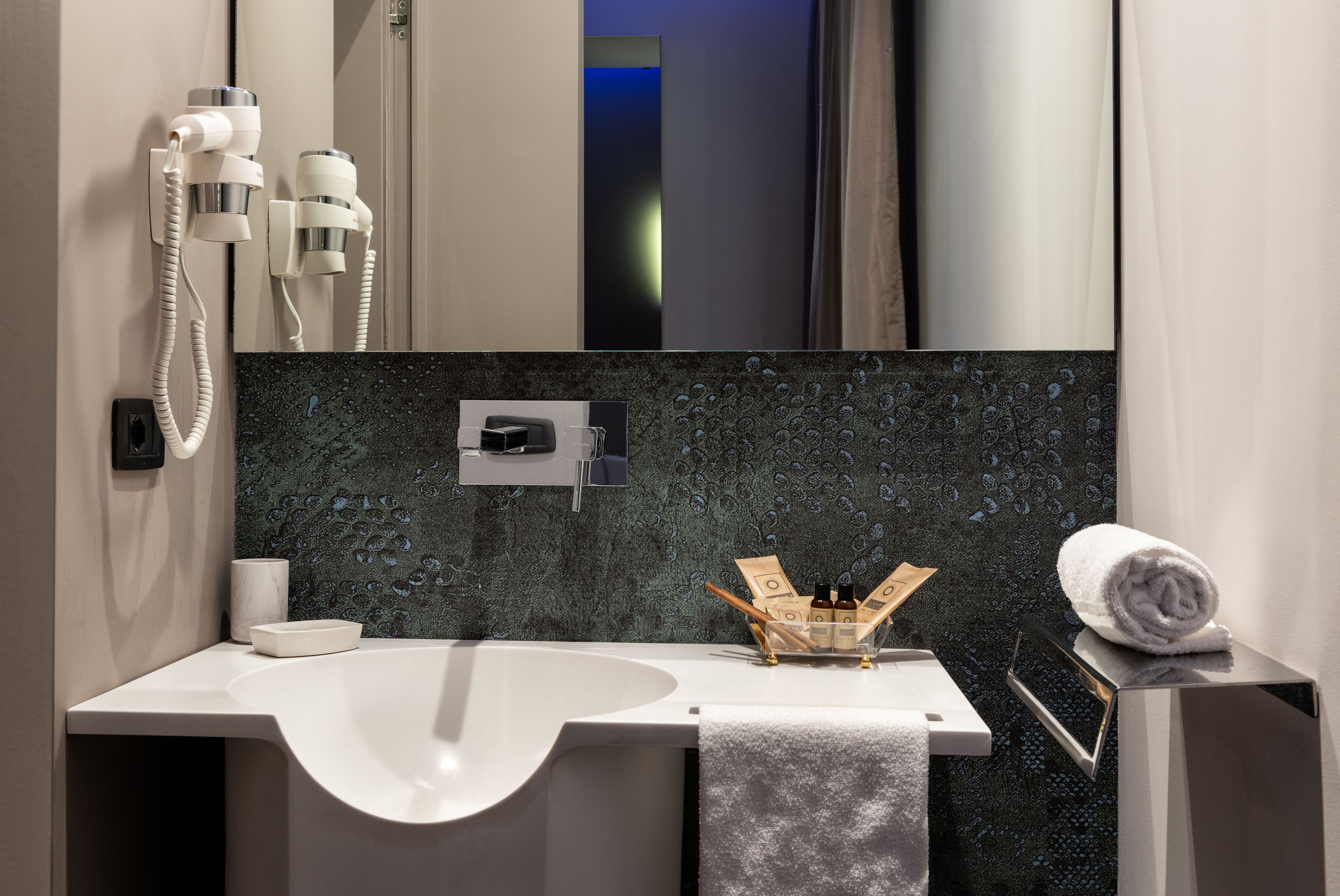 A modern bathroom with a white sink, a mirror, a hairdryer, and a towel on a stand.