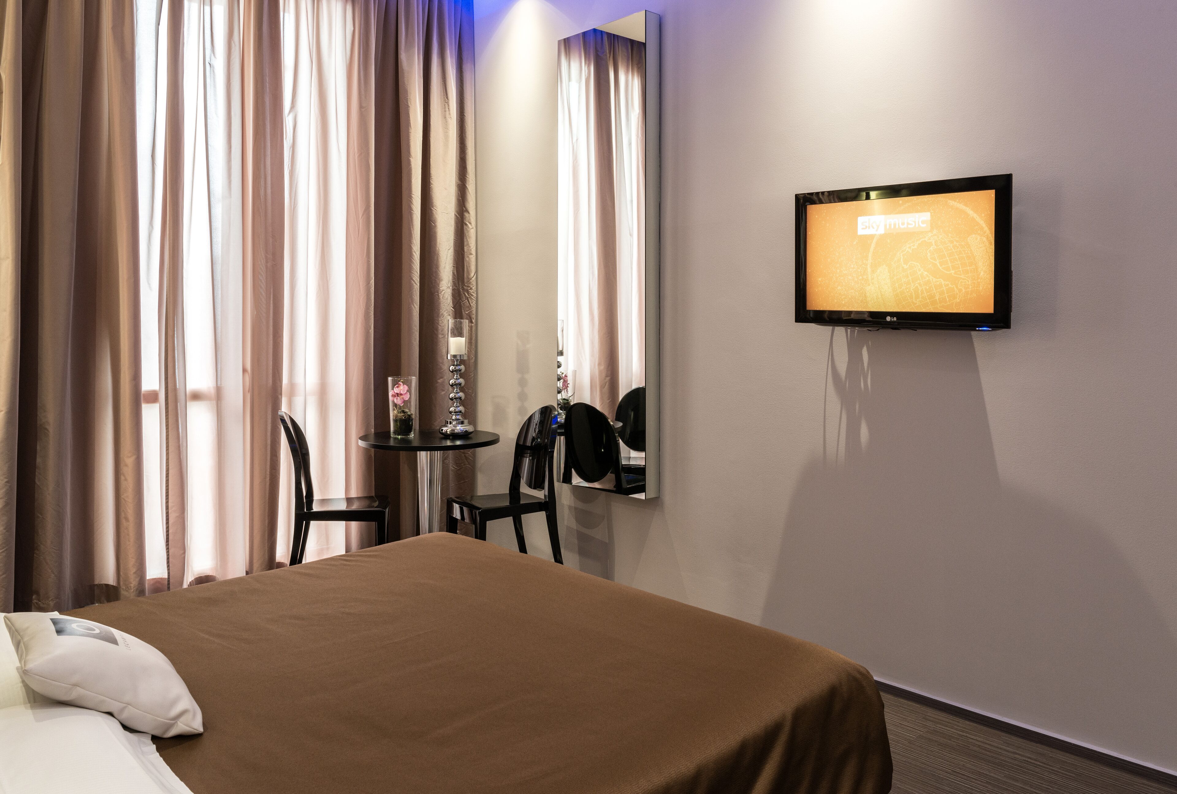 Habitación de hotel con cama, televisor montado en la pared, una mesita con una silla y una ventana con cortinas.