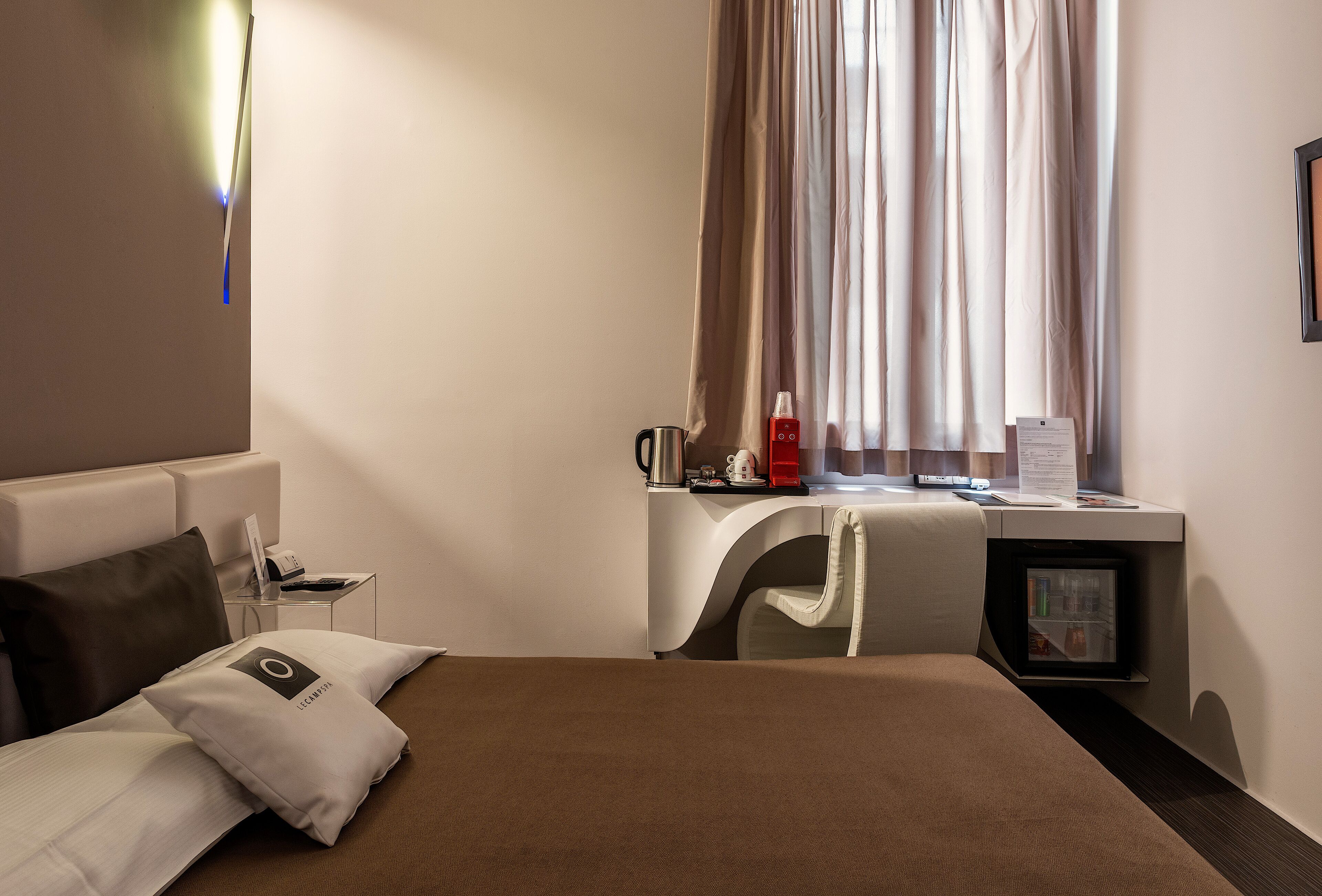 A hotel room with a bed, a desk, a chair, a coffee machine, and a small refrigerator.
