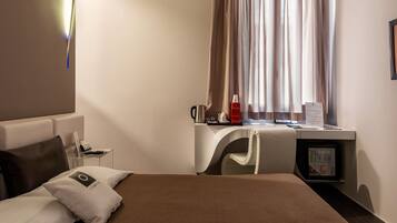 A hotel room with a bed, a desk, a chair, a coffee machine, and a small refrigerator.