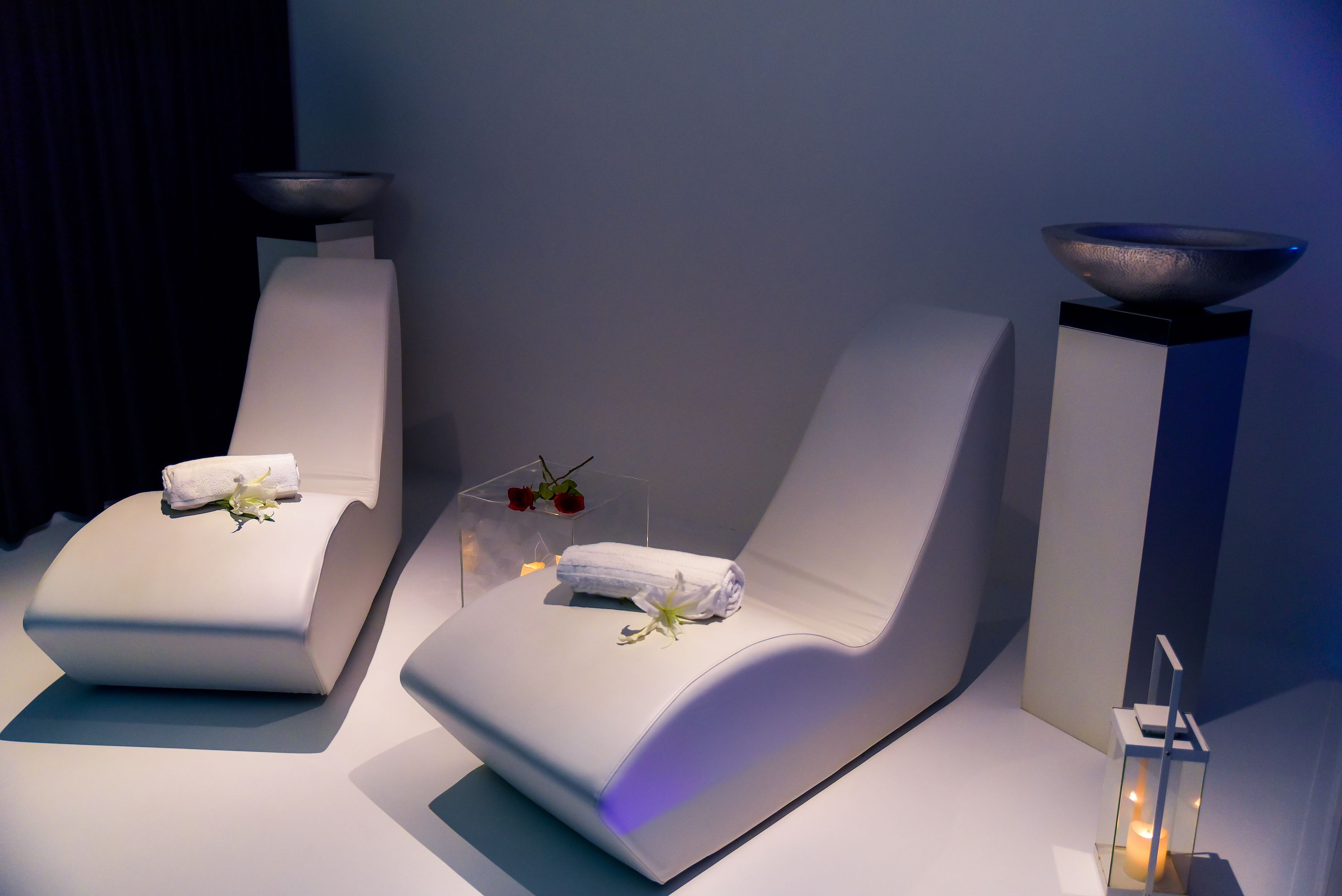 Two white lounge chairs with towels and flowers, a bowl on a pedestal, and a candle in a glass container.