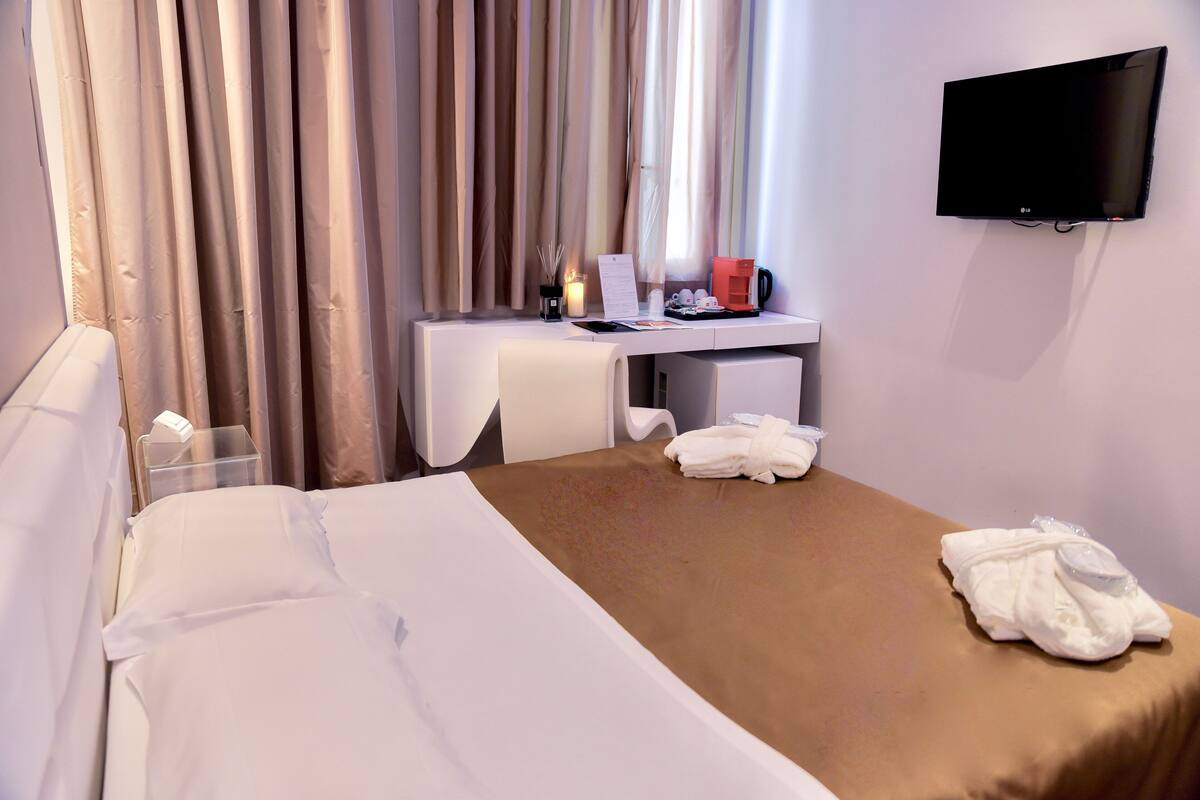 A hotel room with a bed, a desk with a computer, a TV, and a window with curtains.