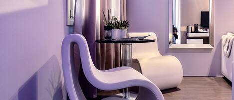 A modern hotel room with a curved white chair, a small table with a plant, a mirror, and a bed.