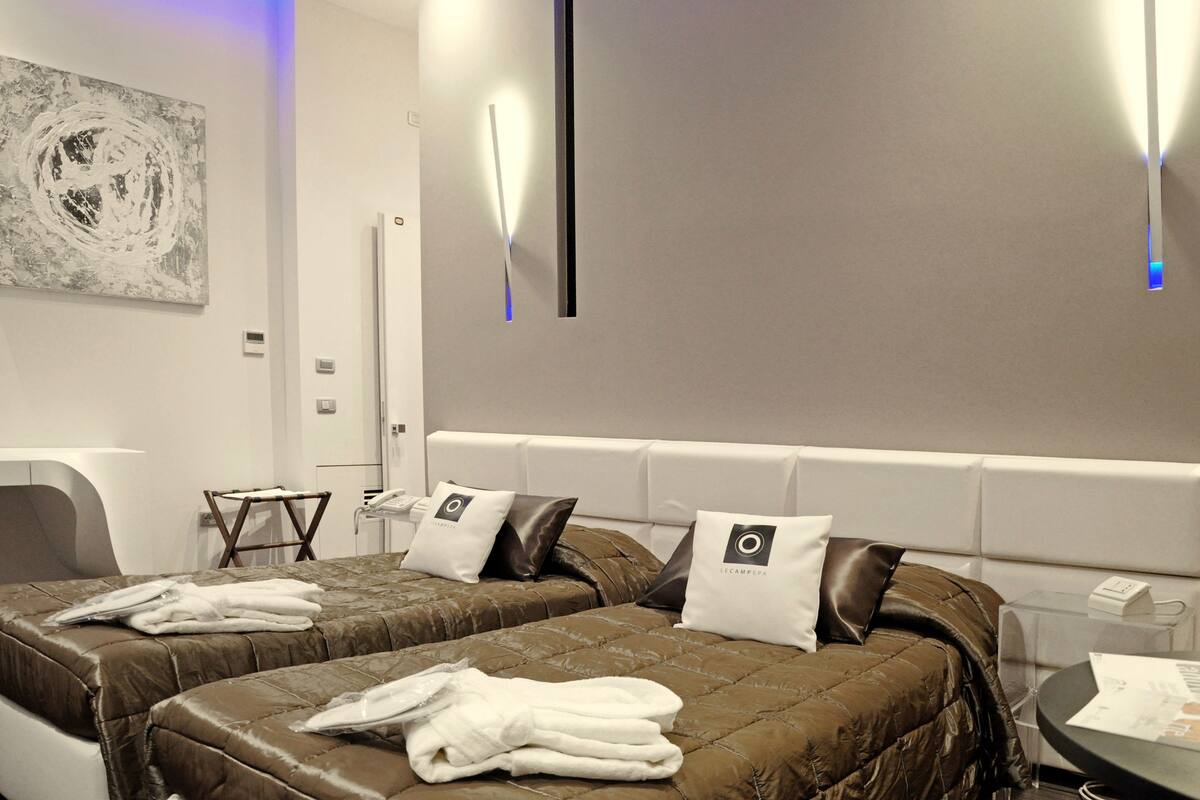 A hotel room with two beds, each with a brown quilted bedspread and white towels. There is a white wall with a modern abstract painting and a wall-mounted light fixture.