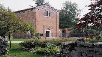 Um edifício de tijolos com uma grande janela em arco central e uma porta pequena, rodeado por vegetação e muros de pedra.