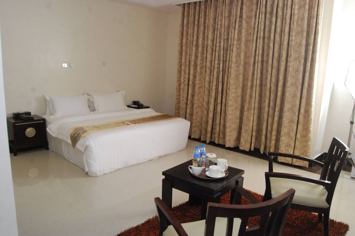 A hotel room with a bed, bedside table, and a small table with cups and bottles.