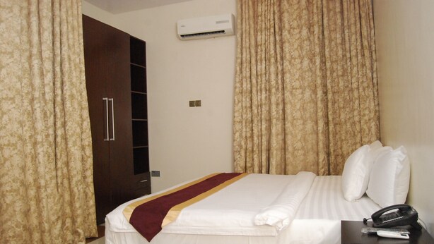 A hotel room with a bed, curtains, an air conditioning unit, a bedside table with a phone, and a wardrobe.