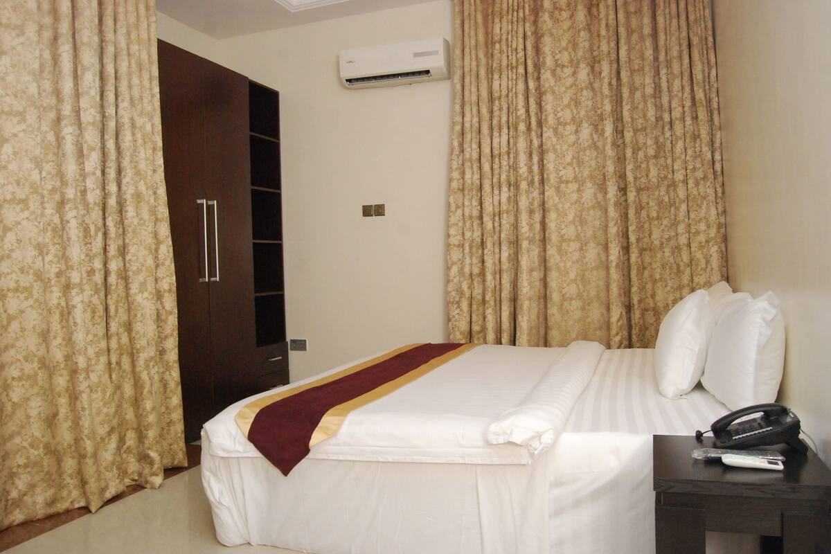 A hotel room with a bed, curtains, an air conditioning unit, a bedside table with a phone, and a wardrobe.