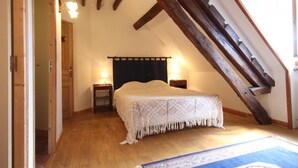 Een slaapkamer met een schuine kap, houten balken, een bed met een gebloemd deken, twee bedlampen en een blauw tapijt met een patroon.