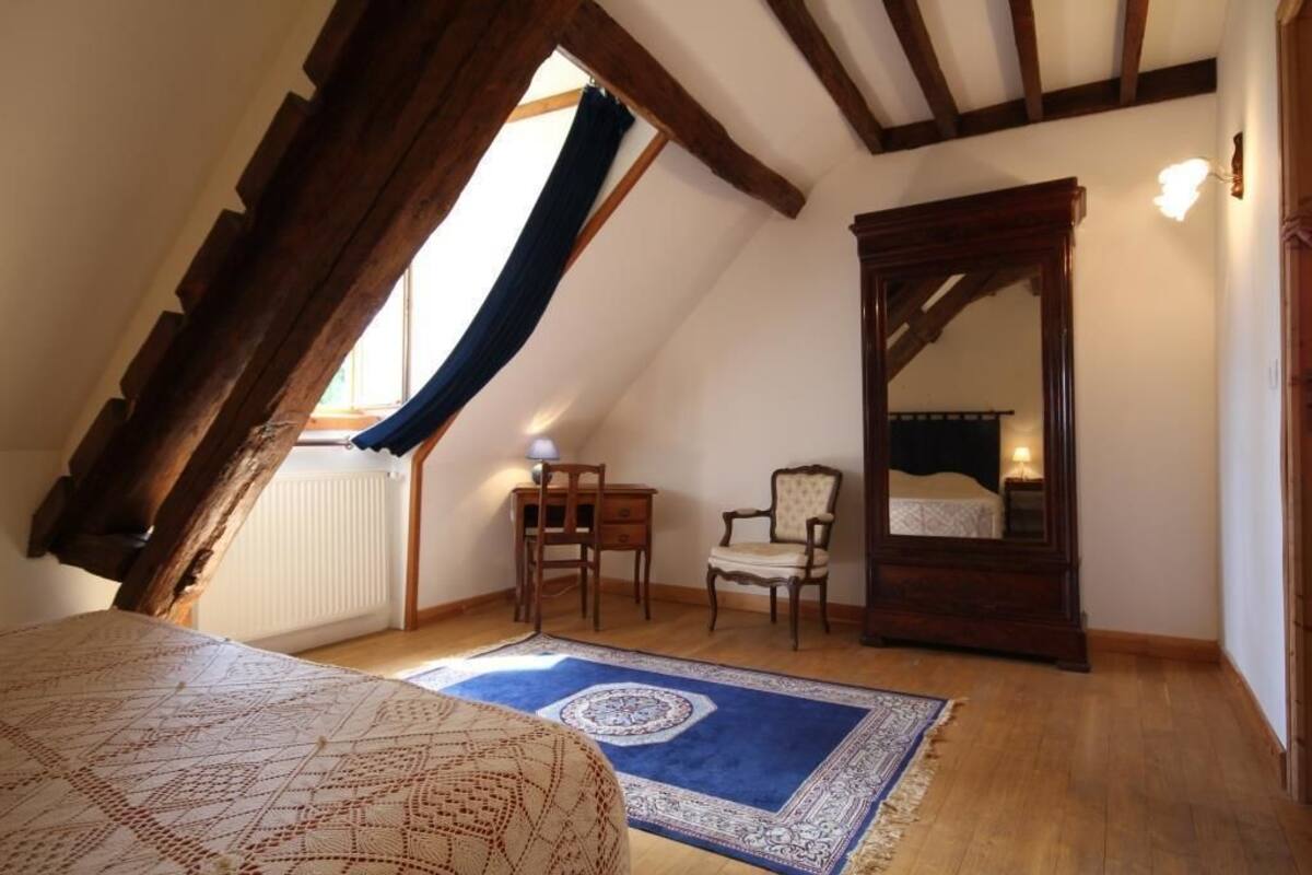 A bedroom with a sloped ceiling, a bed with a patterned bedspread, a blue and white rug, a wooden chair, a nightstand, and a mirror above the bed.