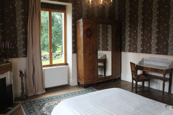A bedroom with a large bed, a wooden wardrobe, a desk, and a window with a view of greenery.