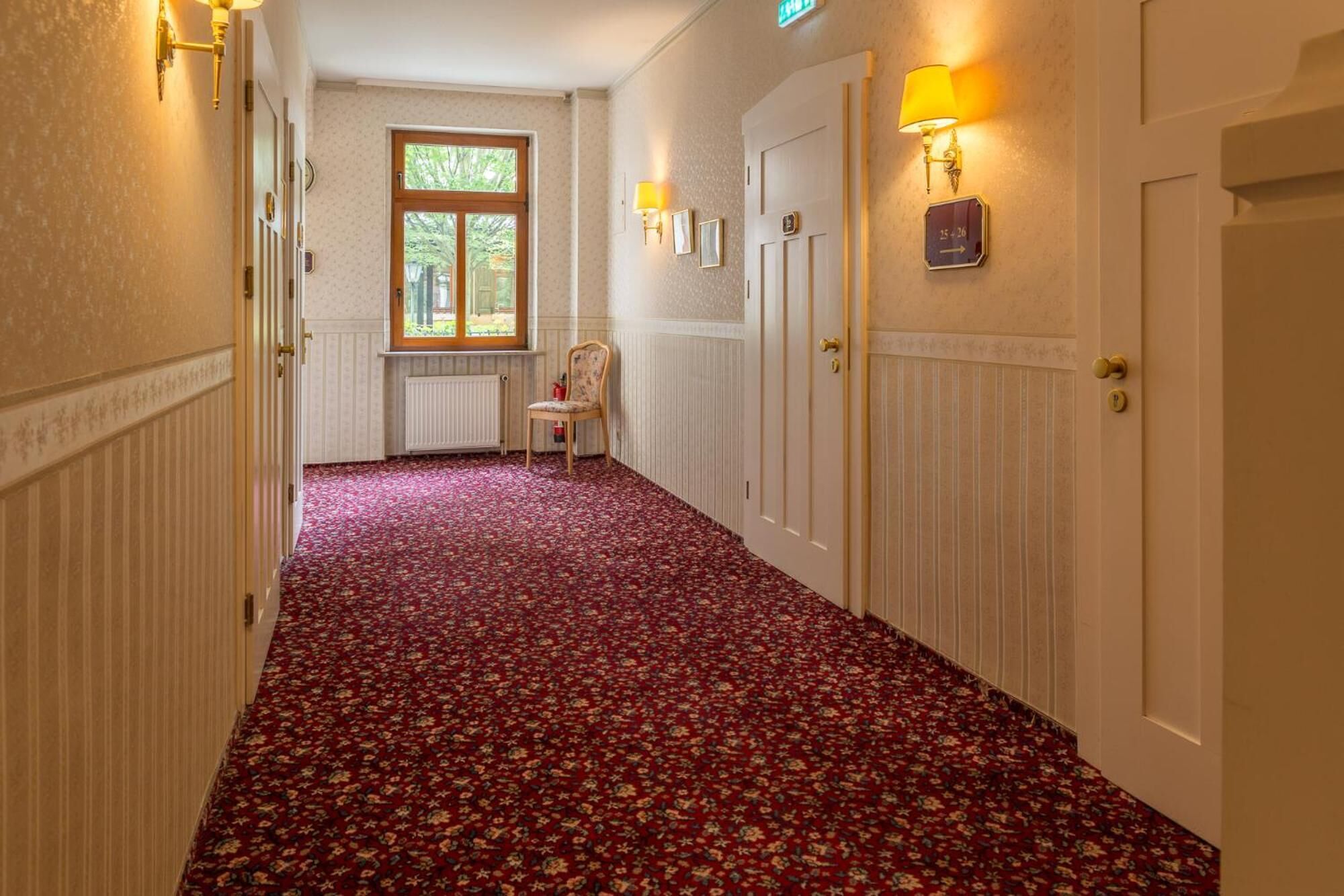 Un pasillo con alfombra estampada, una ventana, una silla y puertas con apliques.