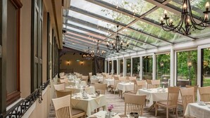 Un espace repas aménagé dans le style d’une véranda, avec des tables dressées pour le service, de grandes fenêtres et un plafond en verre.