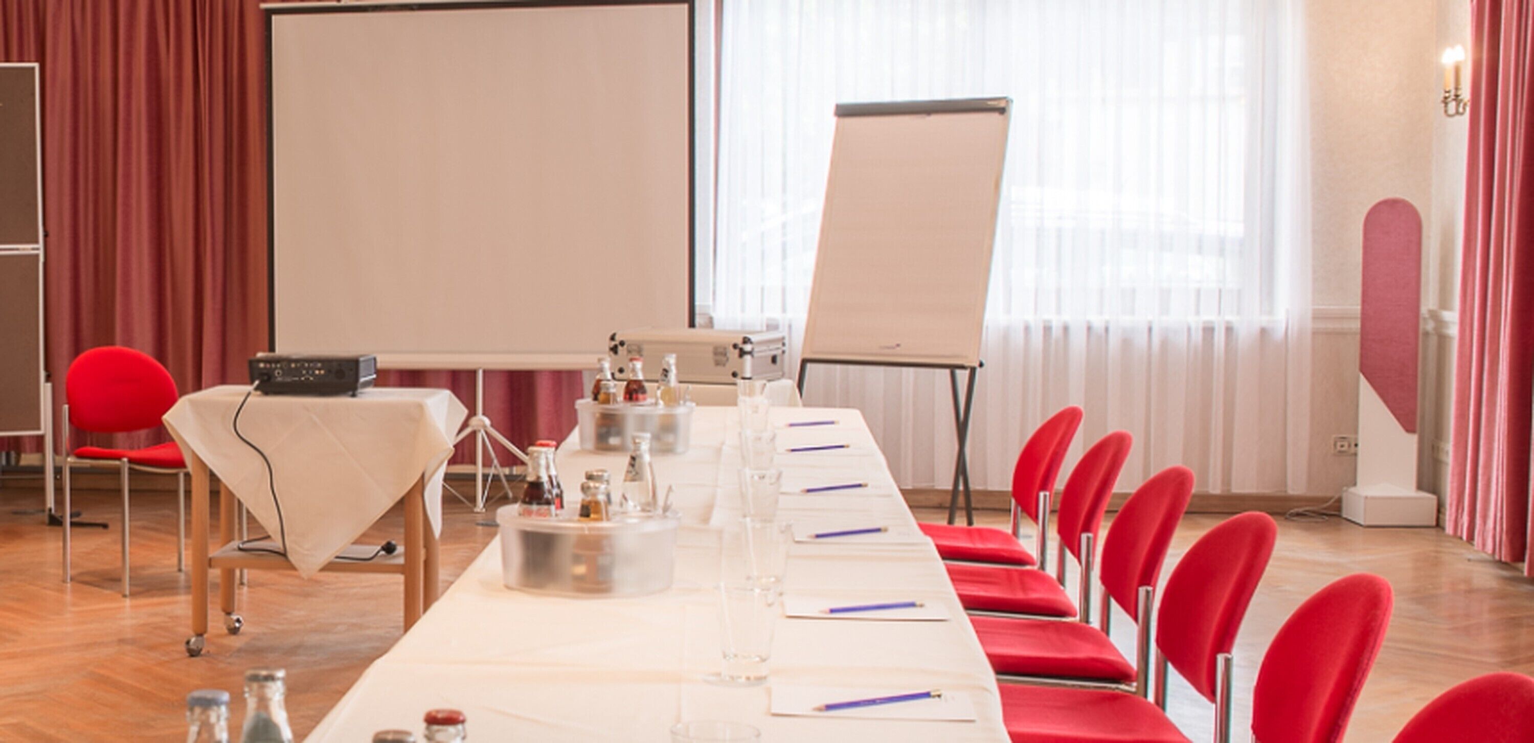 A conference room with a long table, red chairs, a projector screen, and a flip chart.
