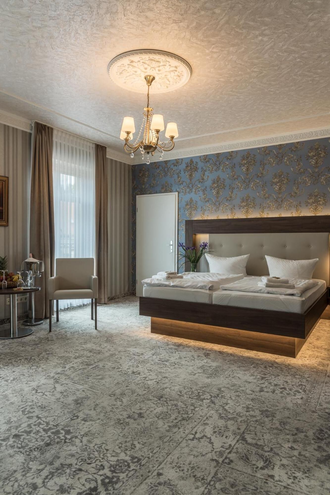 A bedroom with a large bed, a chandelier, a desk with a chair, and a window with curtains.