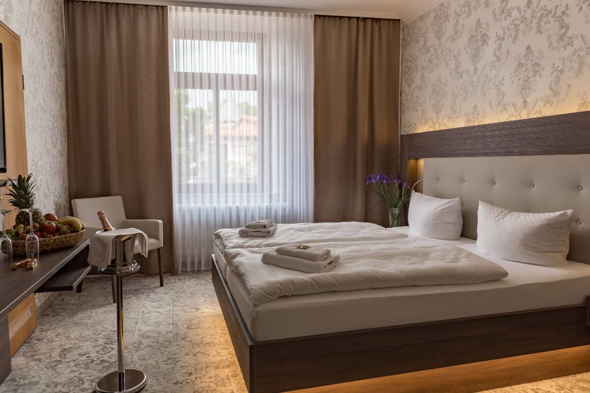 Habitación de hotel con una cama grande, una mesita de noche con frutero, una silla y vistas al exterior a través de cortinas translúcidas.