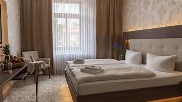 A hotel room with a large bed, a bedside table with a fruit bowl, a chair, and a view of the outdoors through sheer curtains.