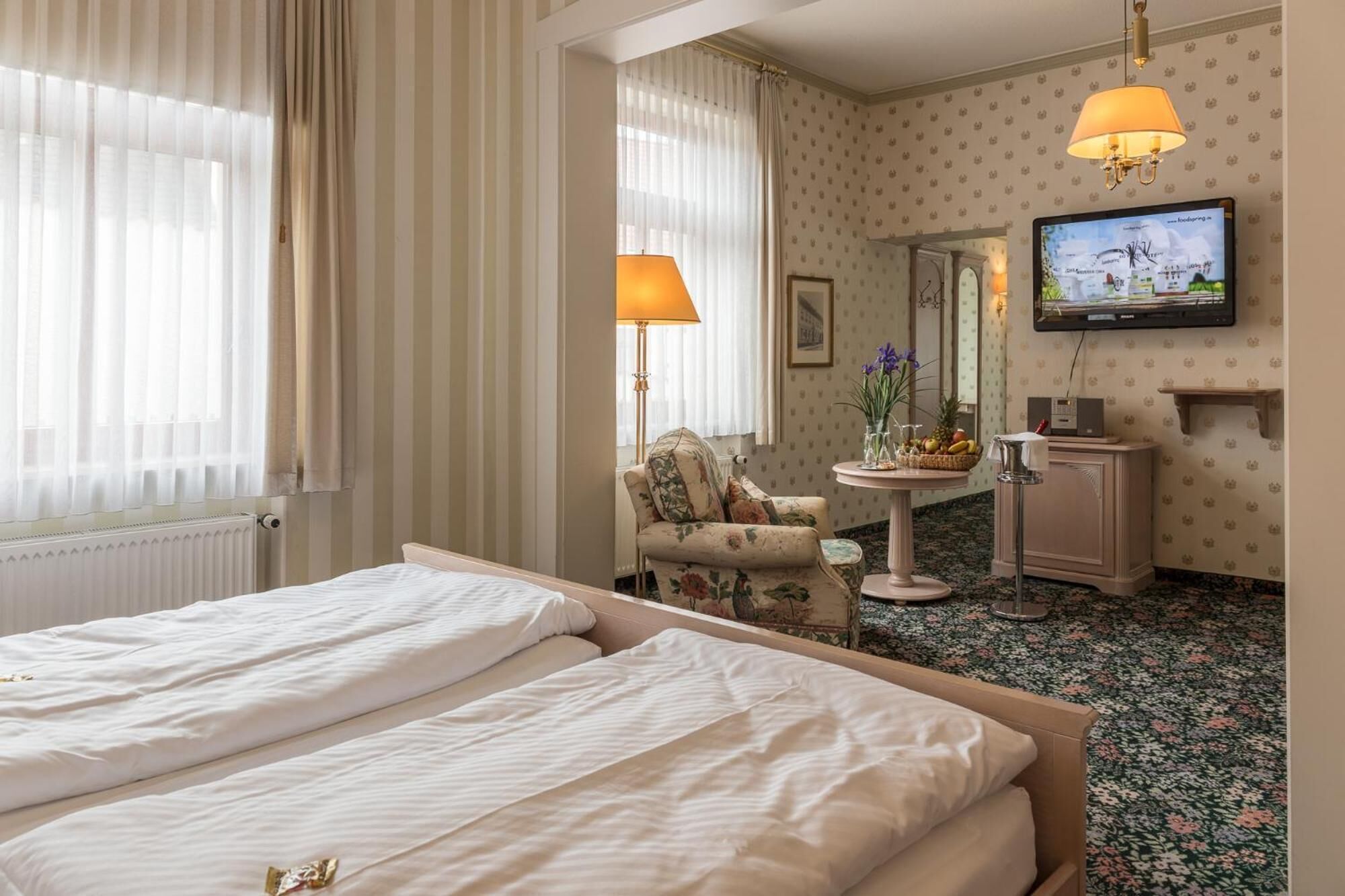 Een hotelkamer met twee bedden, een stoel, een klein tafeltje met fruit, een televisie en een spiegel.