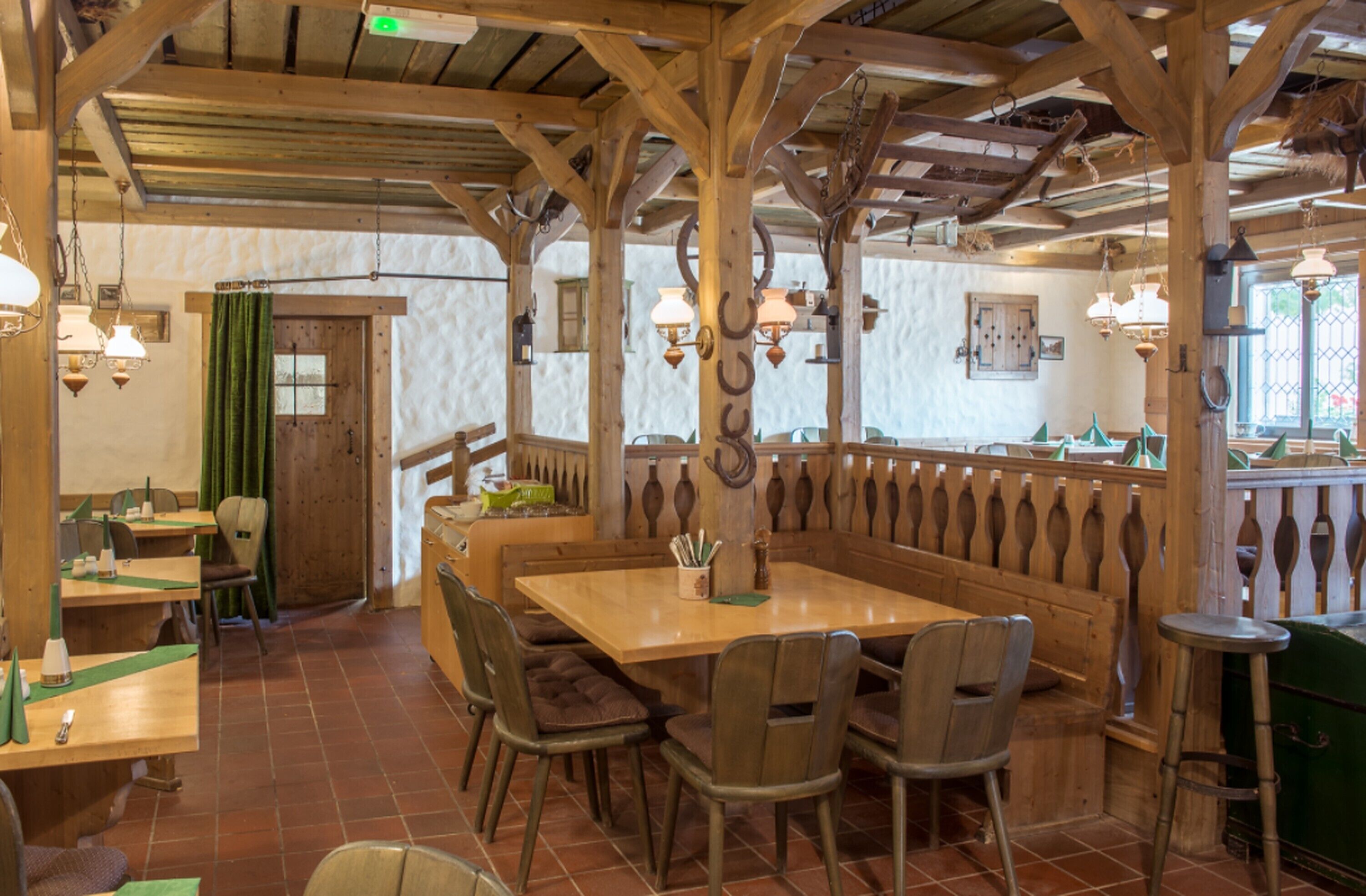 A rustic wooden interior with high ceilings and exposed beams, featuring a central wooden table surrounded by chairs, a green curtain, and hanging lantern-style lights.
