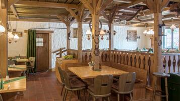 Een rustiek houten interieur met hoge plafonds en zichtbare balken, met een houten tafel in het midden omringd door stoelen, een groen gordijn en hangende lantaarnlampen.
