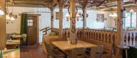 A rustic wooden interior with high ceilings and exposed beams, featuring a central wooden table surrounded by chairs, a green curtain, and hanging lantern-style lights.