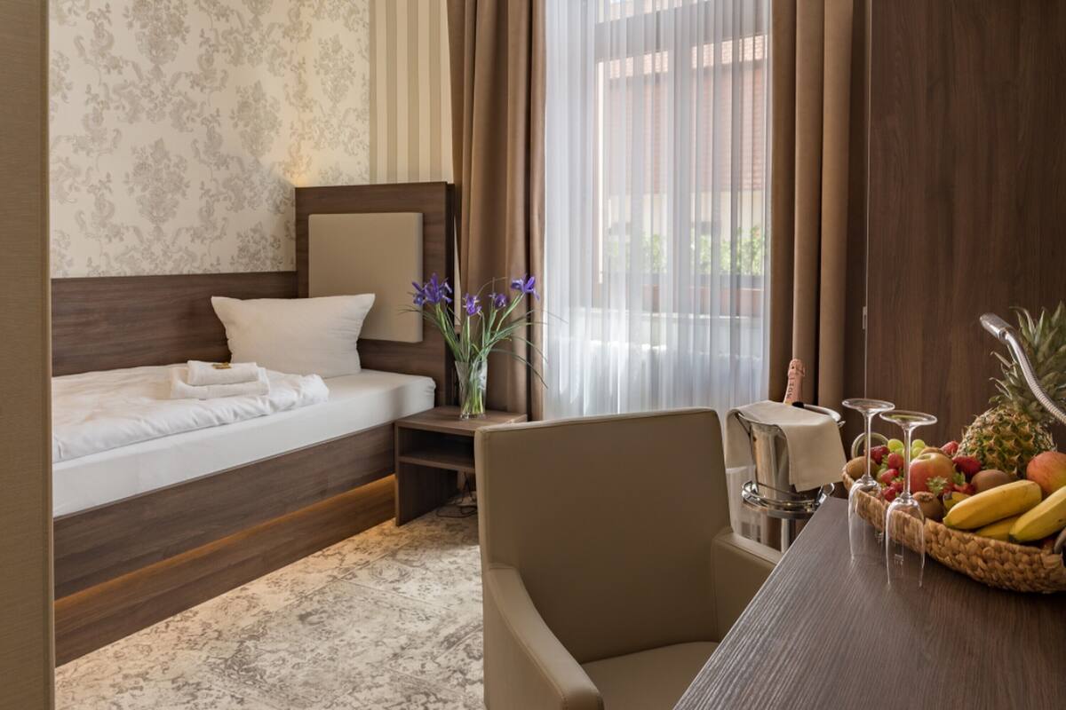 Habitación de hotel con cama, una silla, una mesa con fruta y vasos, y una ventana con cortinas.