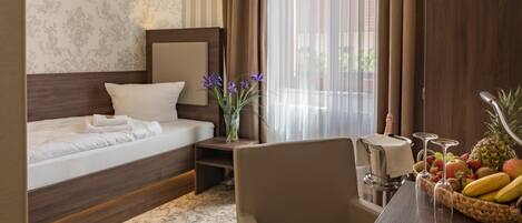 A hotel room with a bed, a chair, a table with fruit and glasses, and a window with curtains.