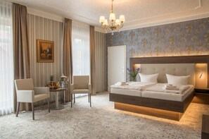 Ein Hotelzimmer mit einem großen Bett, einer Sitzecke mit Stühlen, einem kleinen Tisch mit Obst und einer Vase sowie einem Bild an der Wand.