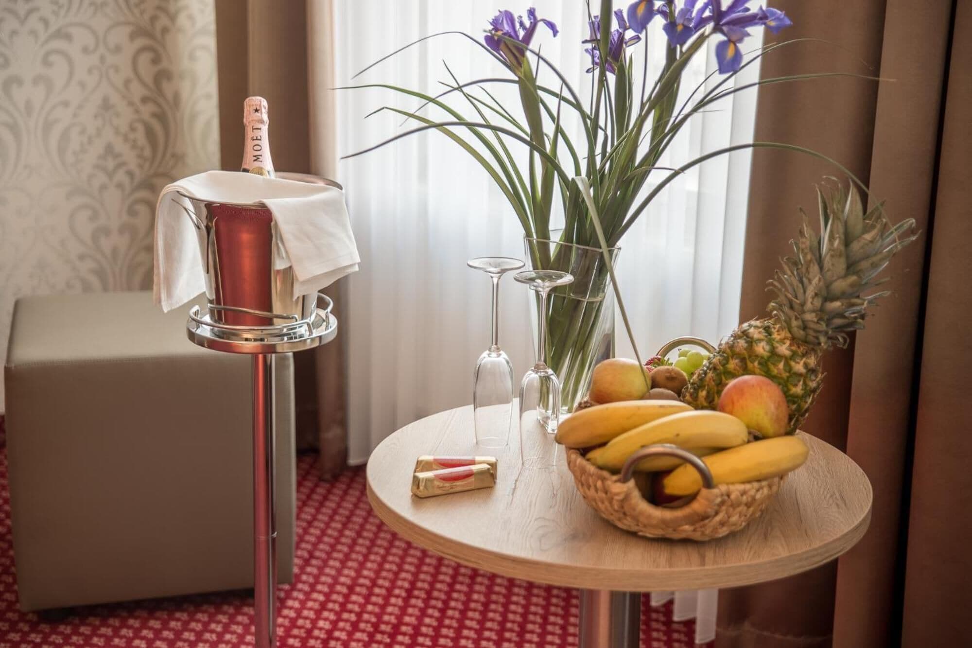 A hotel room with a round wooden table, a minibar with a bottle of Moët champagne, a vase with purple flowers, a fruit basket, and a pineapple.