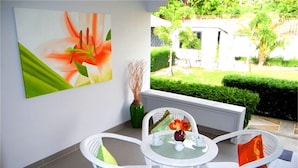 Un pequeño patio al aire libre con una mesa redonda y sillas de mimbre blanco, un jarrón con plantas secas y un cuadro grande de flores en la pared.