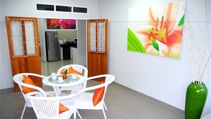 A dining area with white wicker chairs and a round table, a green vase with branches, and a large floral painting on the wall. - Green Leaf 2 Nai Harn 2 bedrooms Villa (Rawai)