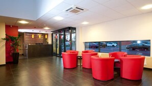 A hotel reception area with a modern design, featuring a reception desk, red seating, and a view of parked cars through large windows. - Hôtel Eurocentre Toulouse Nord 2 Stars (Castelnau-d'Estretefonds)